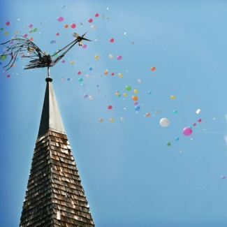 Die Kirchturmspitze mit dem Hahn vor blauem Himmel und fliegenden bunten Luftballons